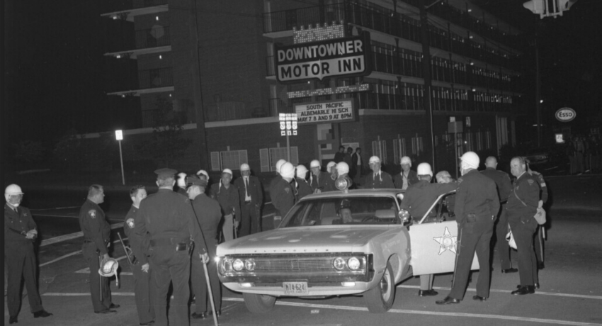 Photograph of law enforcement officers gathering at an intersection in front the Downtowner Motor Inn, 1970