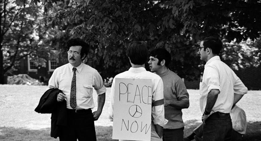 Black and white photograph of Charles Whitebread and law students. One student has a sign on his back which reads: "PEACE NOW"